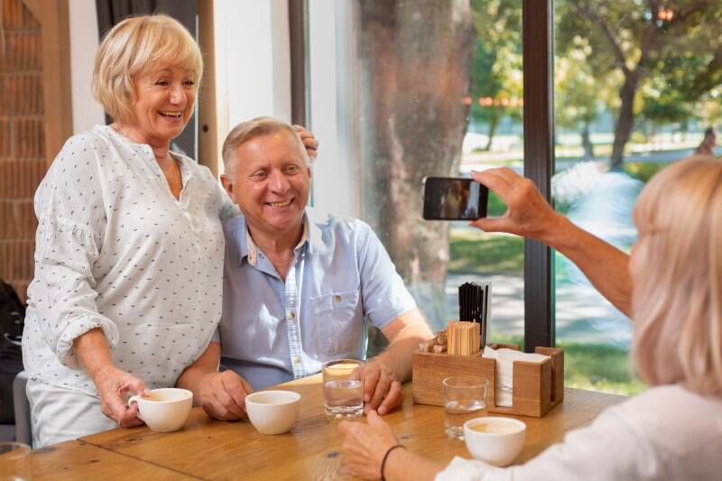 An older couple smiling while a friend takes their photo at a café